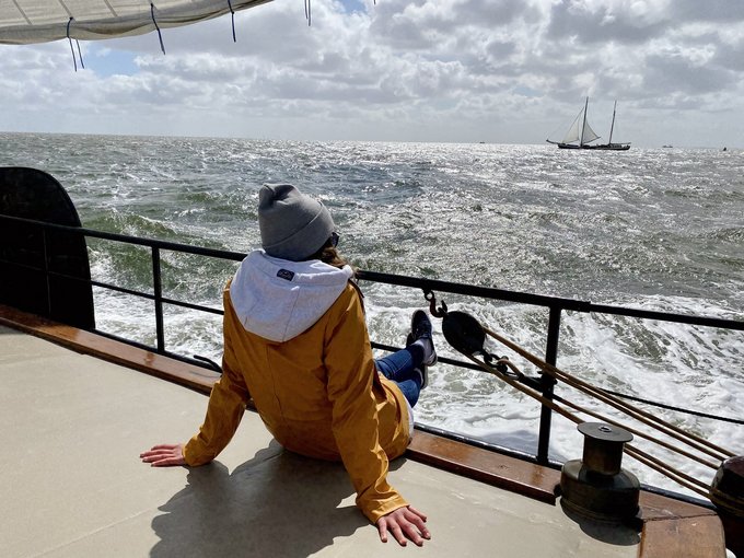 eine Person sitzt entspannt auf einem Boot und blickt über das Meer, am Horizont ein Segelschiff