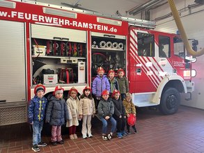 10 Kindergartenkinder mit Feuerwehrhelm vor einem Feuerwehrauto in einer Halle