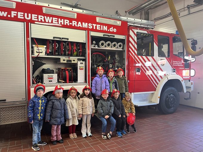 10 Kindergartenkinder mit Feuerwehrhelm vor einem Feuerwehrauto in einer Halle
